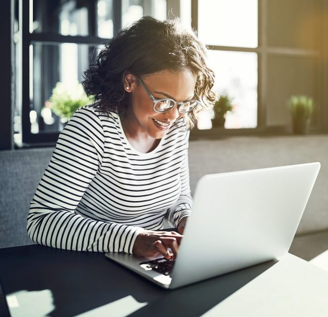 Smiling woman wearing glasses sitting alone at a table browsing the internet with a laptop